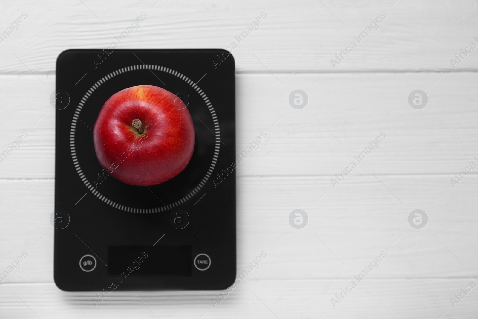 Photo of Digital kitchen scale with ripe red apple on white wooden table, top view. Space for text