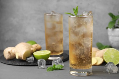 Photo of Glass of tasty ginger ale with ice cubes and ingredients on grey wooden table, space for text