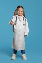 Photo of Little girl in medical uniform showing thumbs up on light blue background