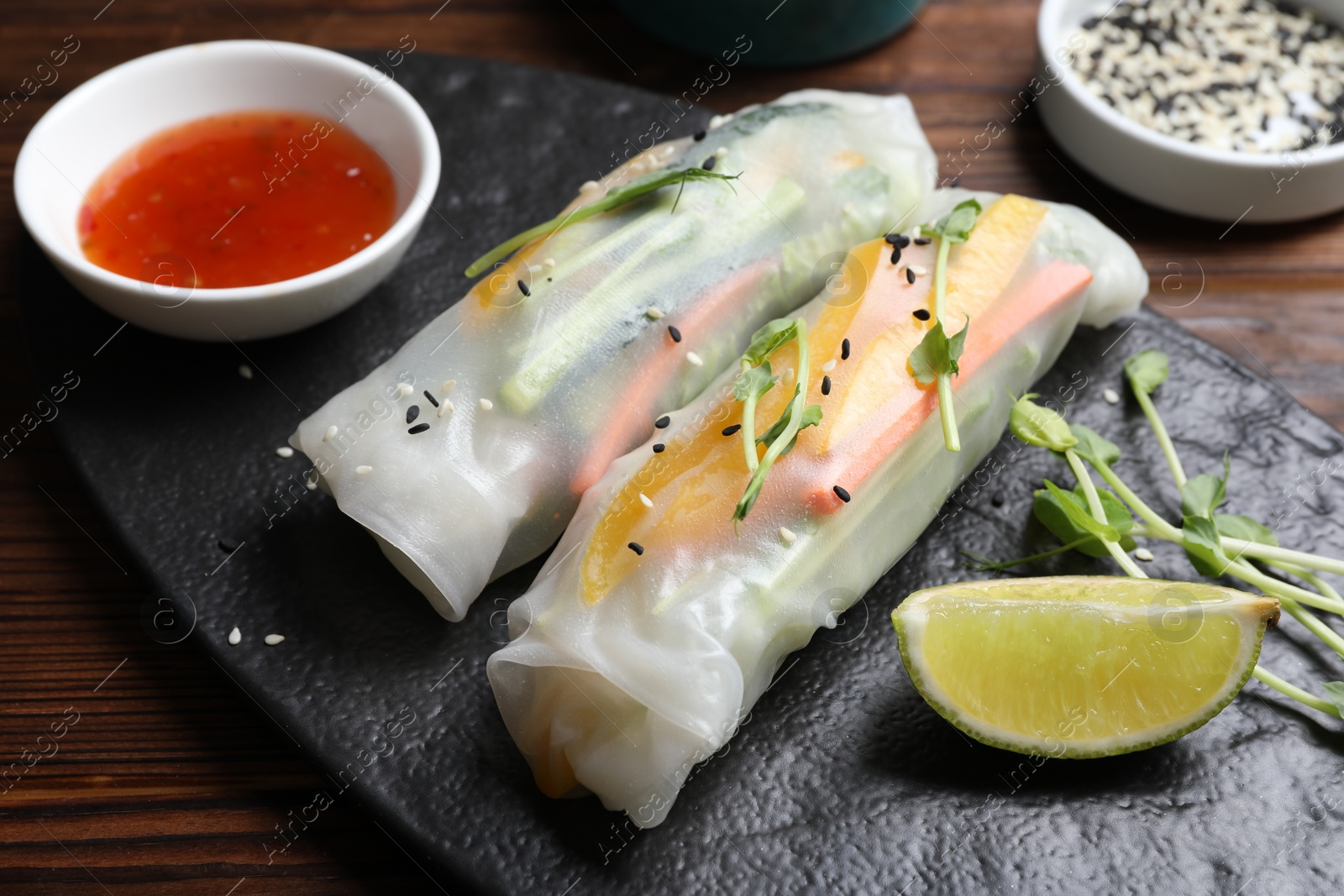 Photo of Tasty spring rolls, sauce, lime, sesame seeds and microgreens on wooden table, closeup