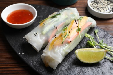 Tasty spring rolls, sauce, lime, sesame seeds and microgreens on wooden table, closeup