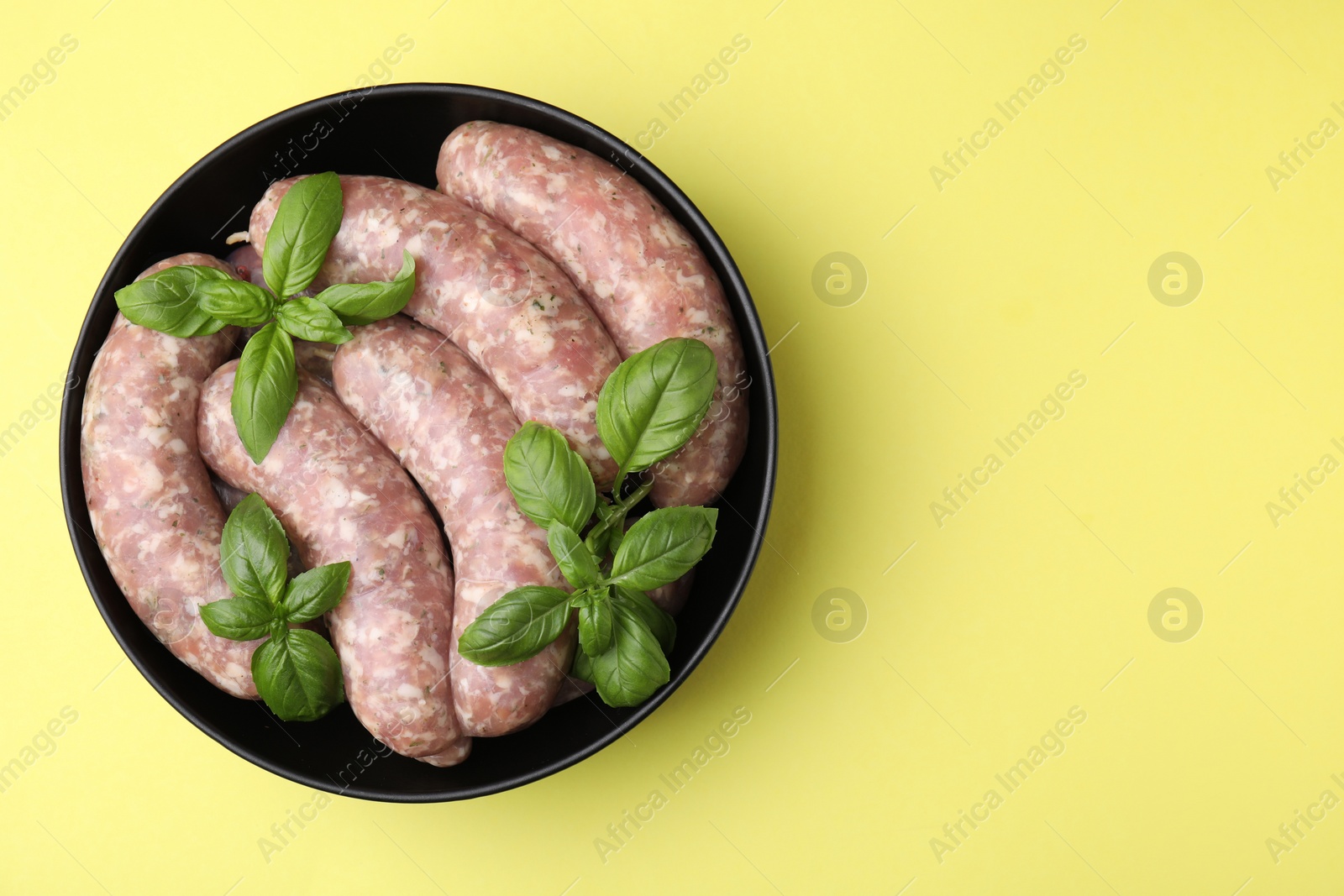 Photo of Raw homemade sausages and basil leaves in bowl on yellow background, top view. Space for text