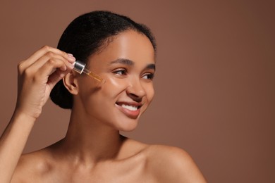 Photo of Smiling woman applying serum onto her face on brown background. Space for text