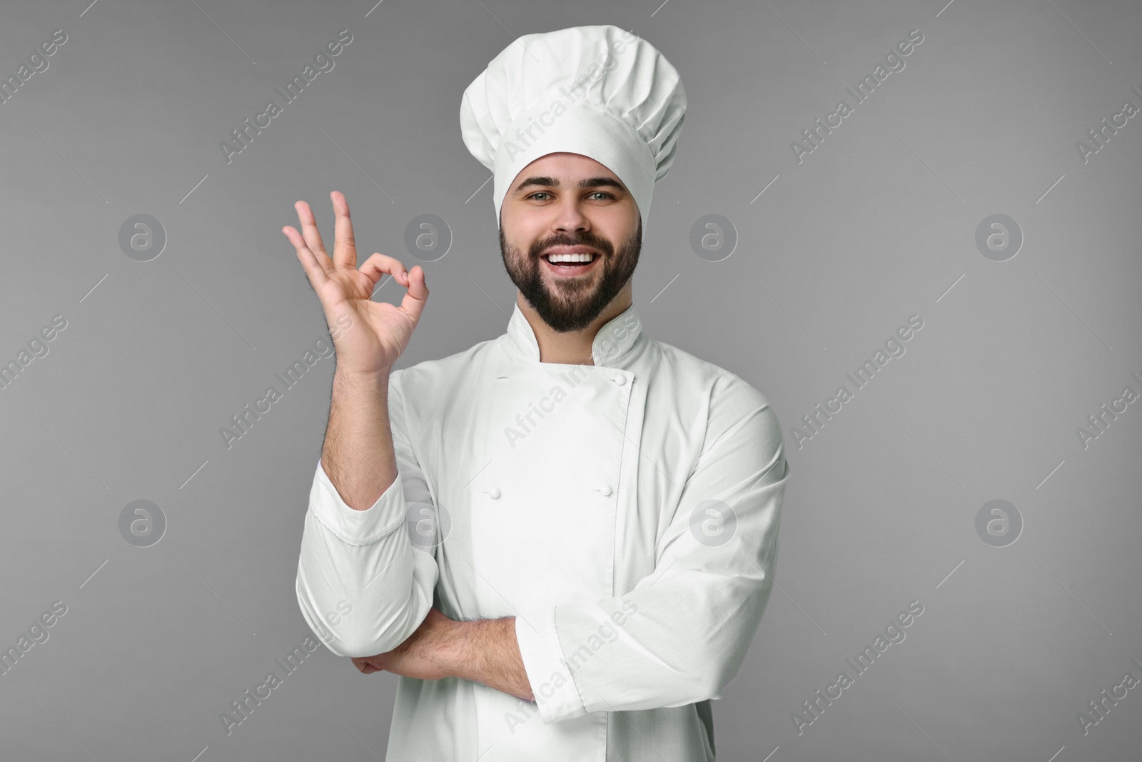 Photo of Happy young chef in uniform showing ok gesture on grey background