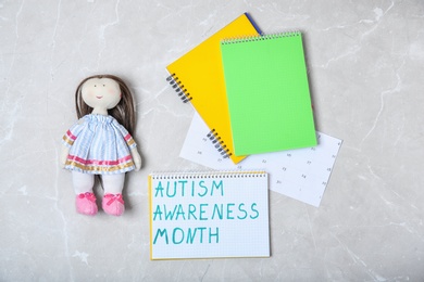 Photo of Notebook with words AUTISM AWARENESS MONTH and doll on grey background, top view