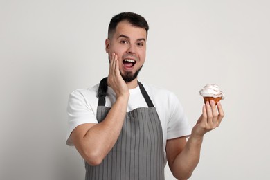 Photo of Happy professional confectioner in apron holding delicious cupcake on light grey background