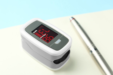 Photo of Modern fingertip pulse oximeter, pen and open notebook on light blue background, closeup