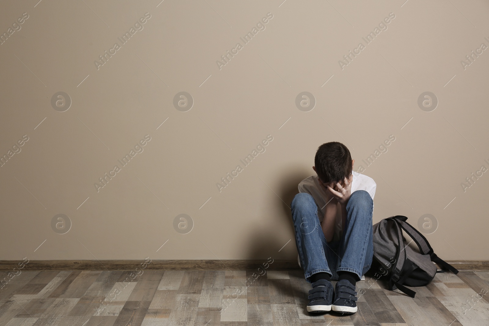 Photo of Upset boy with backpack sitting on floor at color wall. Space for text