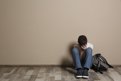 Photo of Upset boy with backpack sitting on floor at color wall. Space for text