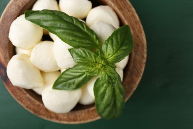 Photo of Tasty mozzarella balls and basil leaves in bowl on green wooden table, top view