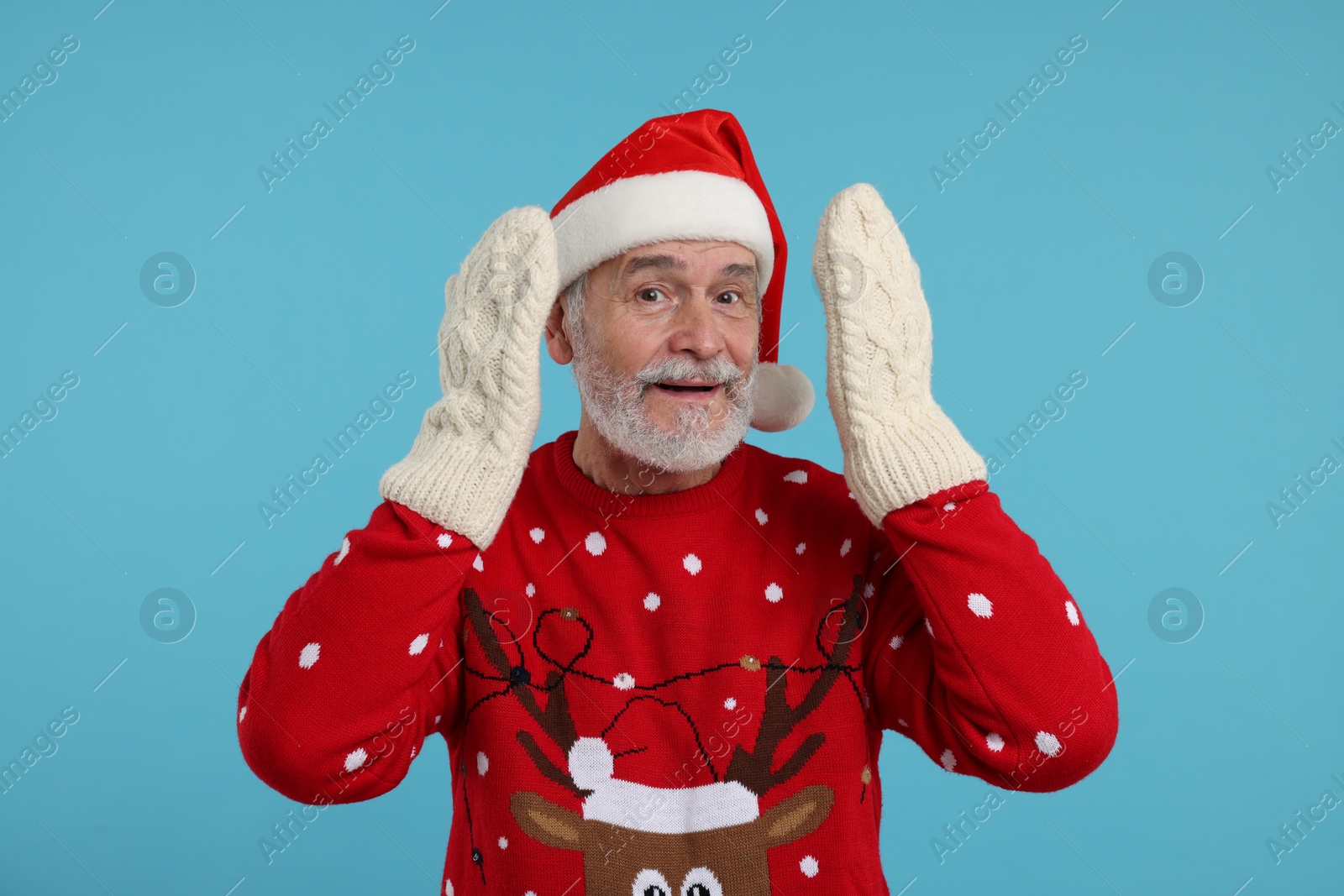 Photo of Senior man in Christmas sweater, Santa hat and knitted mittens on light blue background