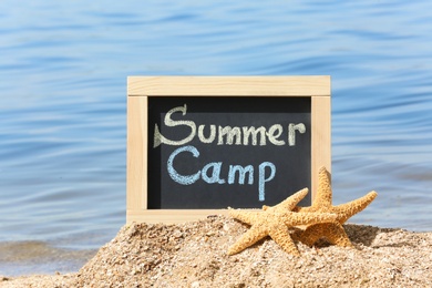 Photo of Little blackboard with text SUMMER CAMP and sea stars on sand near sea