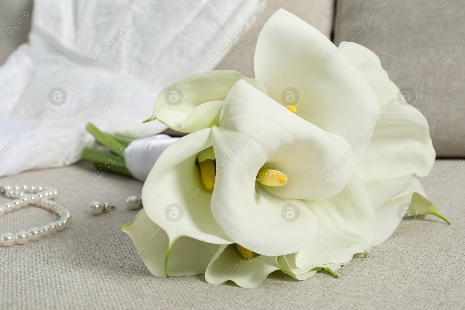 Photo of Beautiful calla lily flowers tied with ribbon, wedding dress and jewelry on sofa