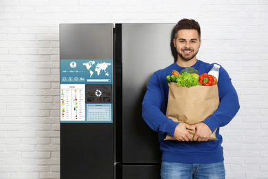 Image of Young man with bag of groceries near smart refrigerator indoors
