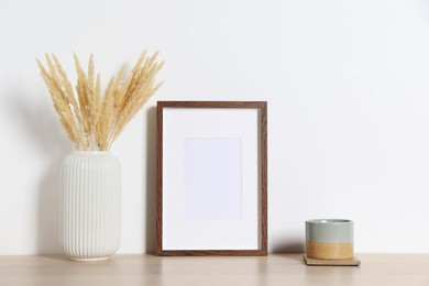 Photo of Empty photo frame, cup and vase with dry decorative spikes on wooden table. Mockup for design