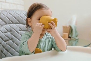 Photo of Cute little baby drinking from cup in high chair indoors