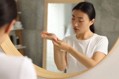 Photo of Suffering from allergy. Young woman scratching her arm near mirror in bathroom