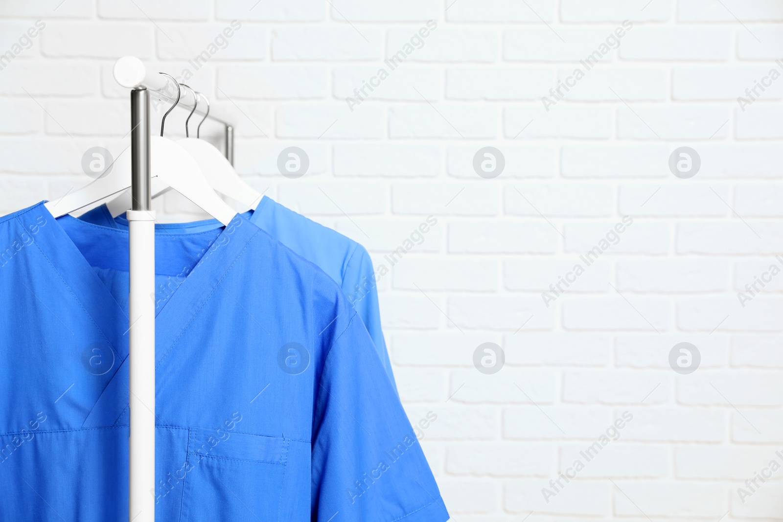 Photo of Medical uniforms hanging on rack near white brick wall. Space for text