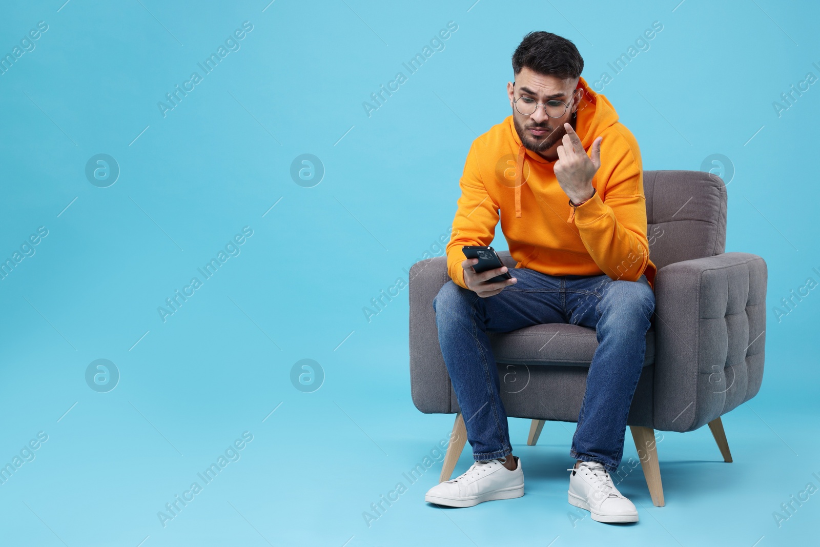 Photo of Emotional young man using smartphone on armchair against light blue background, space for text