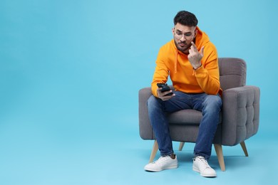 Emotional young man using smartphone on armchair against light blue background, space for text