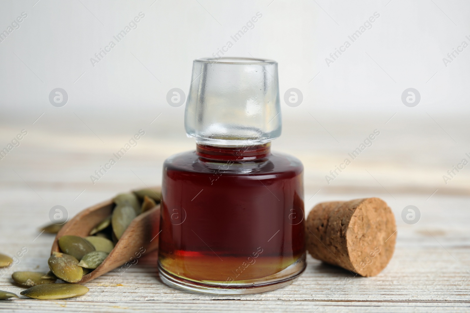 Photo of Glass bottle of oil and scoop with pumpkin seeds on white wooden table, closeup