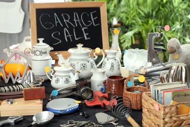 Photo of Blackboard with sign Garage Sale and many different stuff on black wooden table indoors