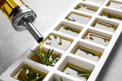 Pouring olive oil into ice cube tray with fresh rosemary at grey table, closeup