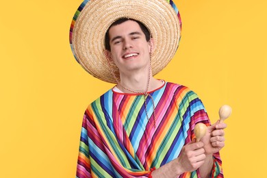 Young man in Mexican sombrero hat and poncho with maracas on yellow background