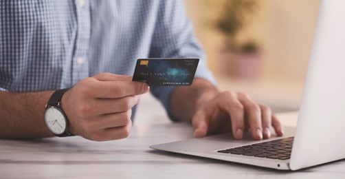 Man with credit card using laptop for online payment at table, closeup. Banner design