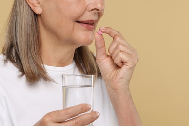 Senior woman with glass of water taking pill on beige background, closeup