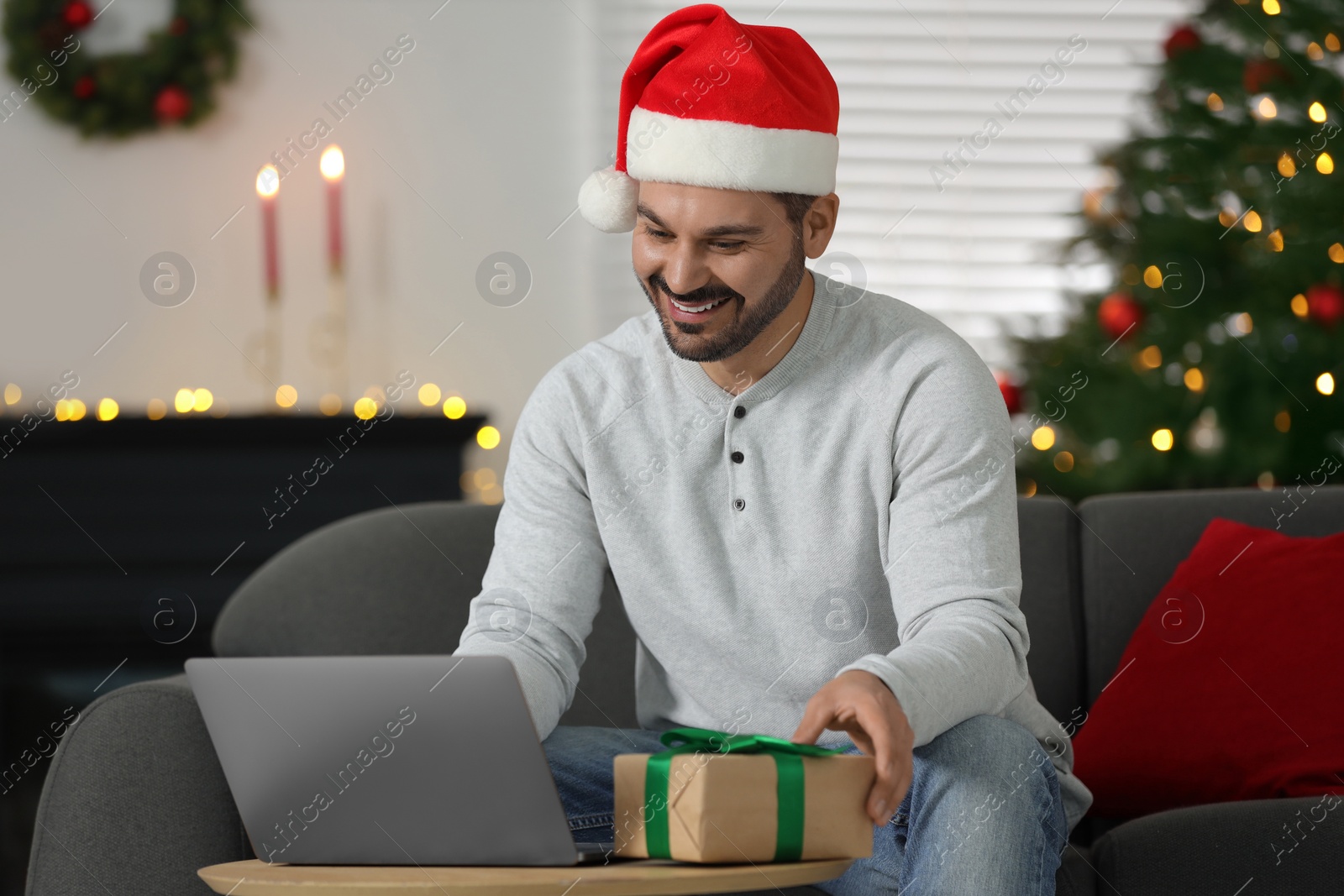 Photo of Celebrating Christmas online with exchanged by mail presents. Happy man in Santa hat with gift box during video call on laptop at home