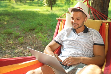 Young man with laptop resting in comfortable hammock at green garden
