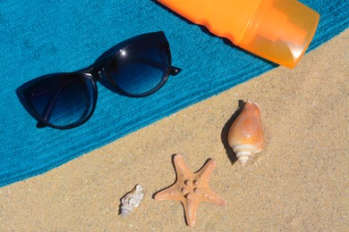 Stylish sunglasses, bottle of sunblock and blue towel on sand, flat lay. Beach accessories