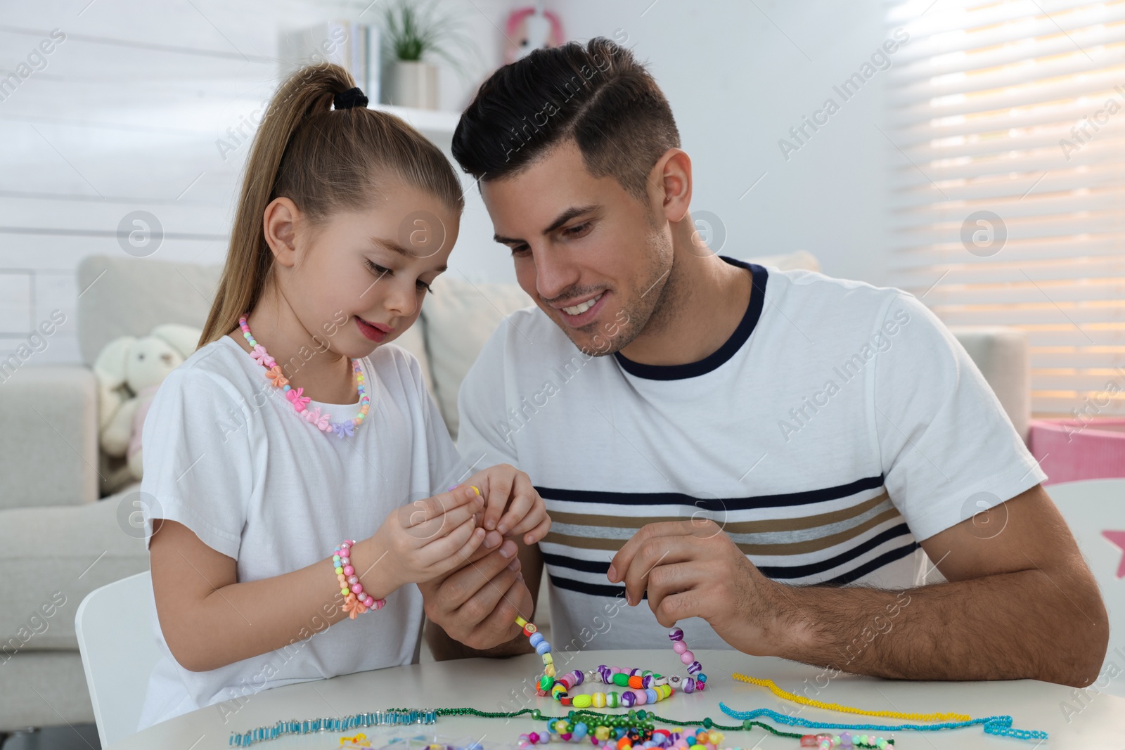 Photo of Happy father with his cute daughter making beaded jewelry at table in room