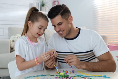 Happy father with his cute daughter making beaded jewelry at table in room