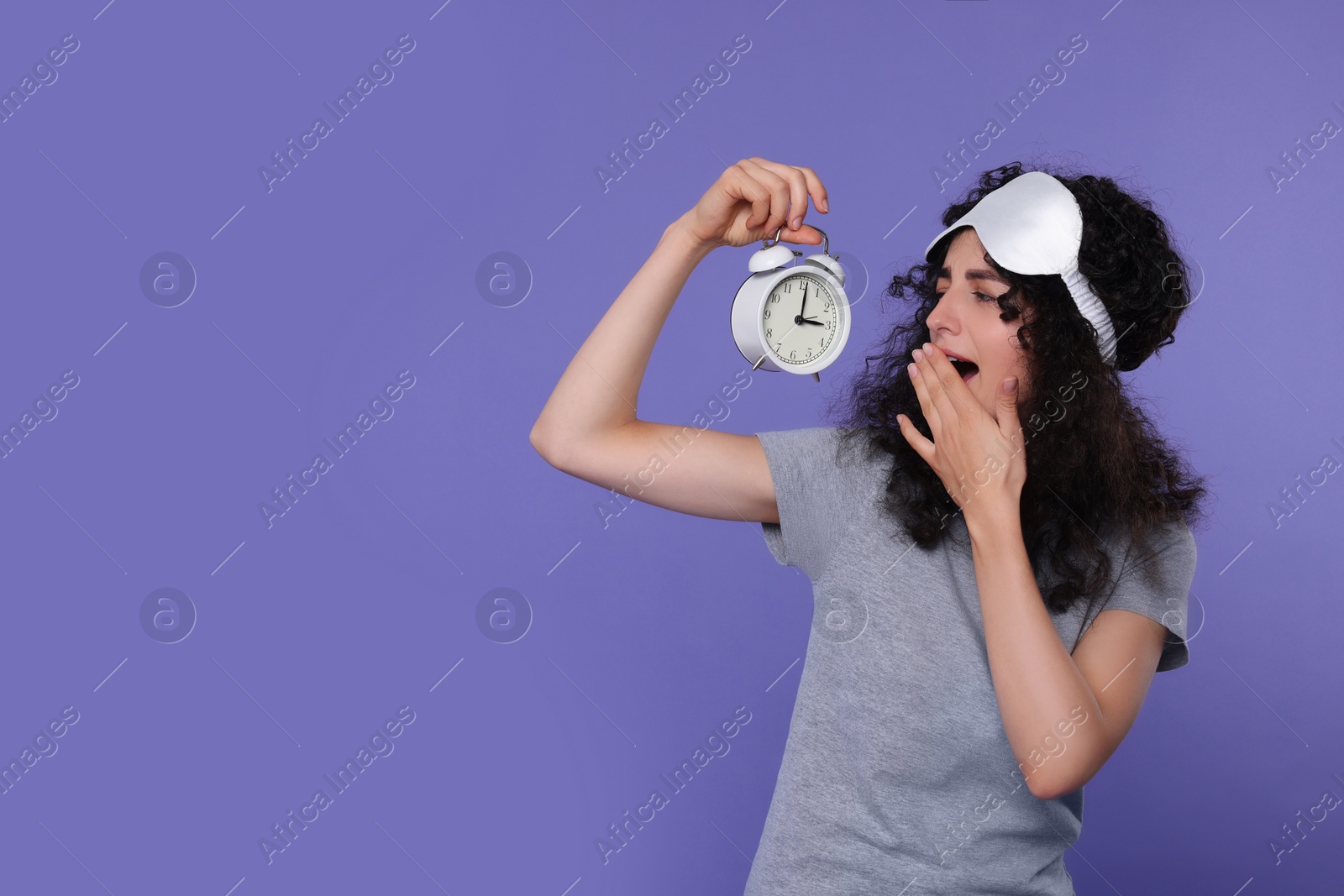 Photo of Tired young woman with sleep mask and alarm clock yawning on purple background, space for text. Insomnia problem