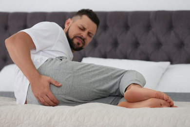 Photo of Man suffering from hemorrhoid on bed at home, focus on hand