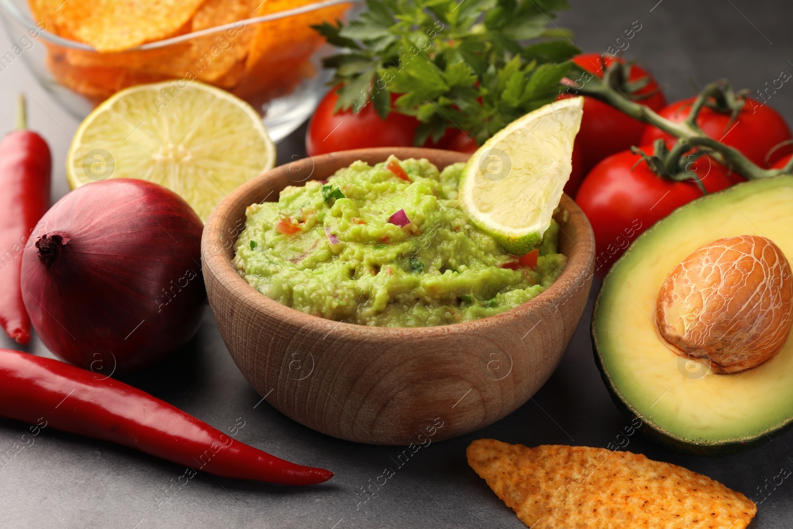 Photo of Bowl of delicious guacamole, nachos chips and ingredients on grey table, closeup