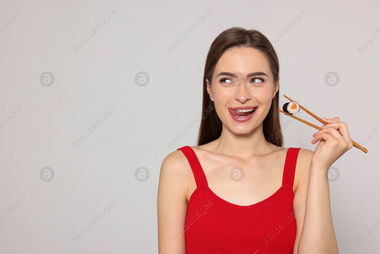 Photo of Happy young woman holding sushi roll with chopsticks on light background. Space for text