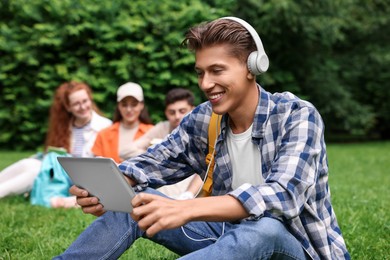 Photo of Students learning together in park. Happy young man with headphones working with tablet on green grass, selective focus