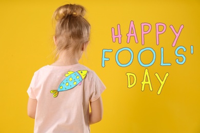 Image of Little girl with paper fish on back against yellow background. Happy fool's day