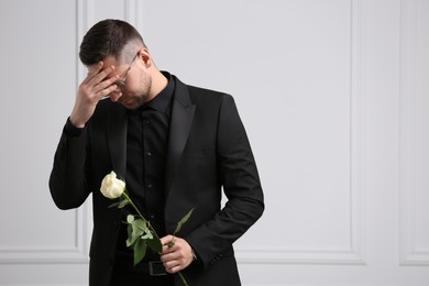 Sad man with rose flower mourning near white wall, space for text. Funeral ceremony