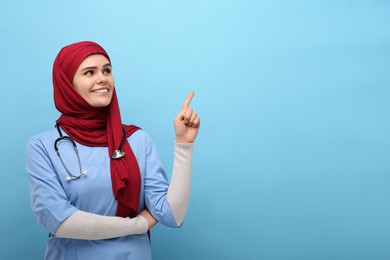 Photo of Muslim woman in hijab and medical uniform pointing at something on light blue background, space for text