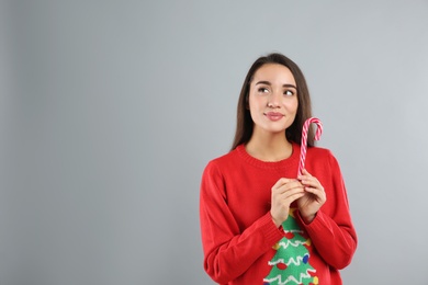 Young woman in Christmas sweater holding candy cane on grey background, space for text