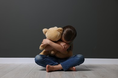 Photo of Child abuse. Upset girl with toy sitting on floor near grey wall