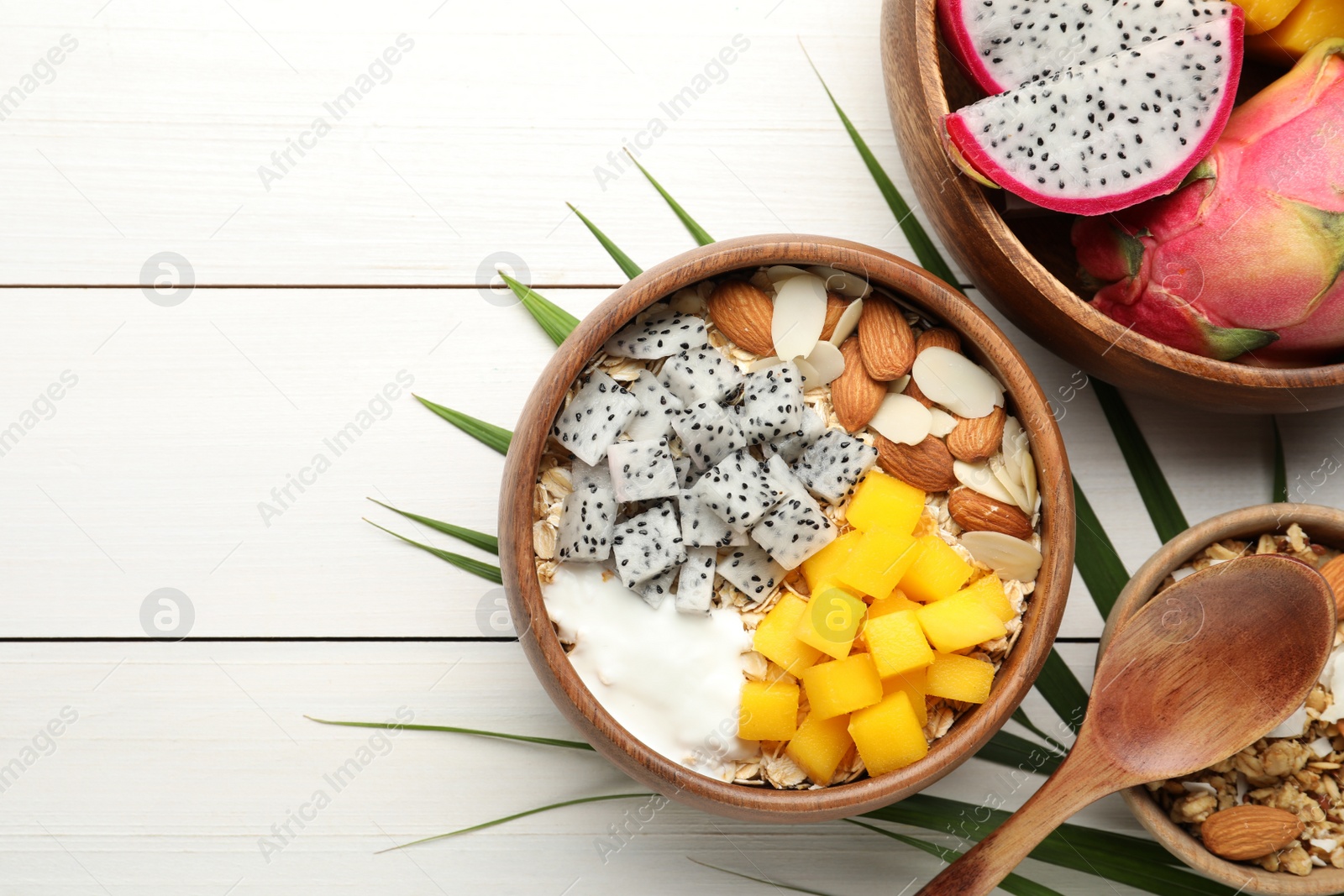 Photo of Bowl of granola with pitahaya, mango, almonds and yogurt on white wooden table, flat lay. Space for text