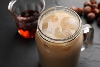 Photo of Mason jar of delicious iced coffee, syrup and hazelnuts on black table, closeup