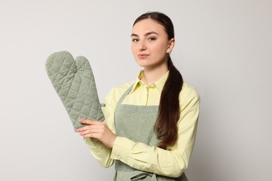 Beautiful young woman in clean apron with pattern and oven glove on light grey background