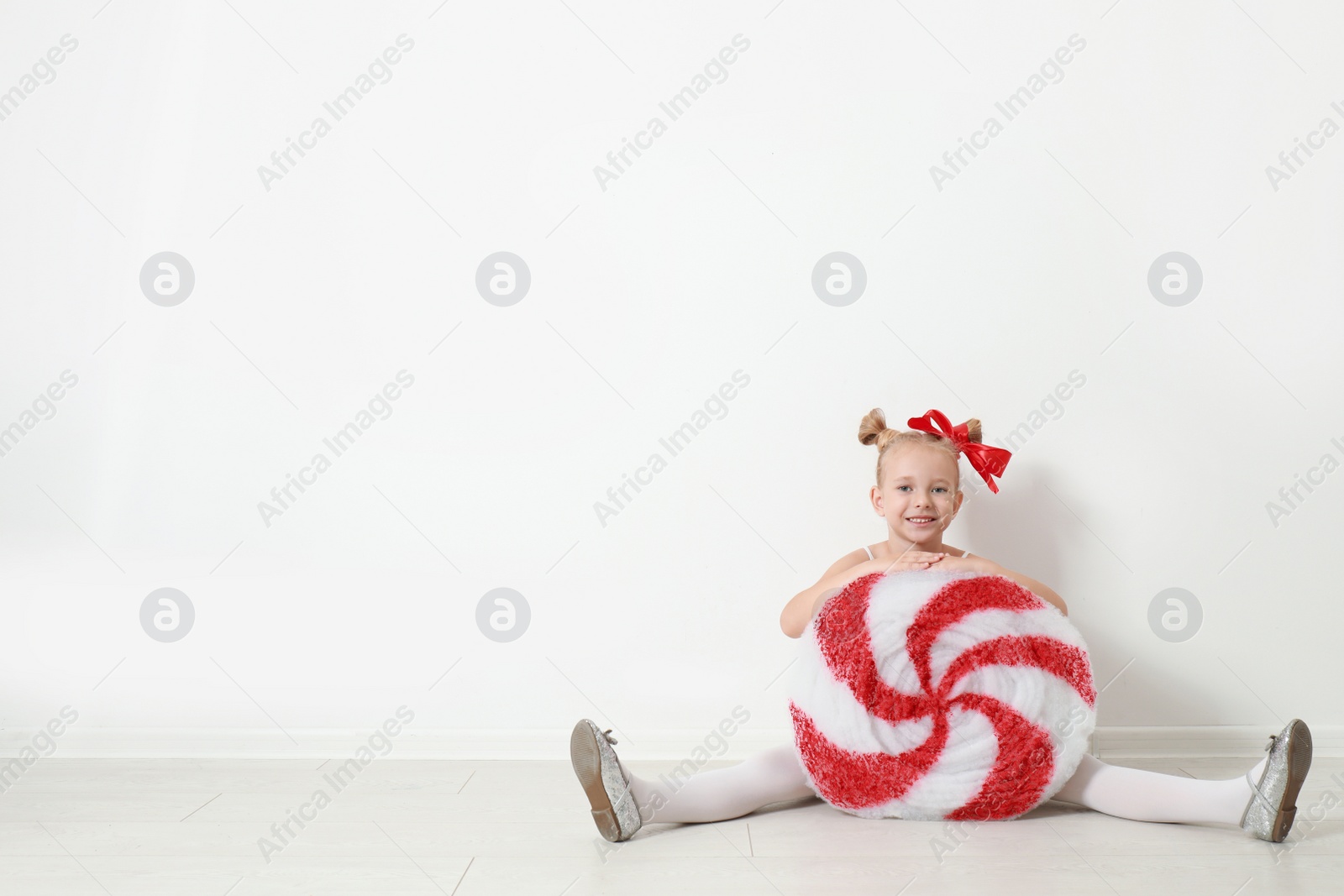 Image of Cute little girl dressed as candy sitting near white wall, space for text. Christmas suit