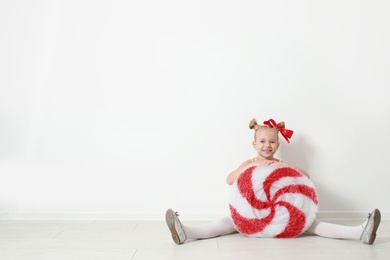 Image of Cute little girl dressed as candy sitting near white wall, space for text. Christmas suit